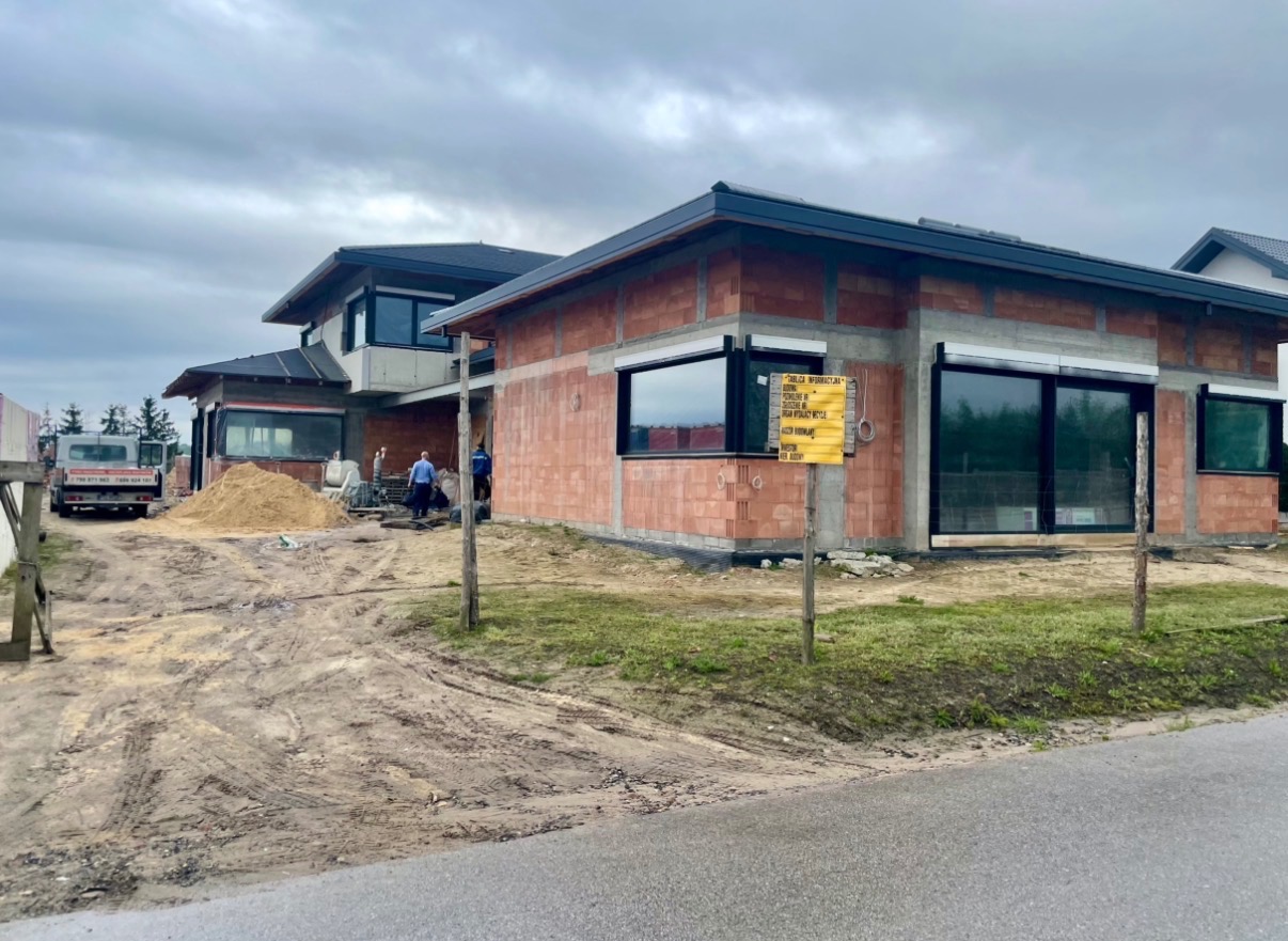 Budowa domu jednorodzinnego w stanie surowym otwartym, widoczne ściany z czerwonej cegły, okna z roletami zewnętrznymi, plac budowy z piaskiem i sprzętem.