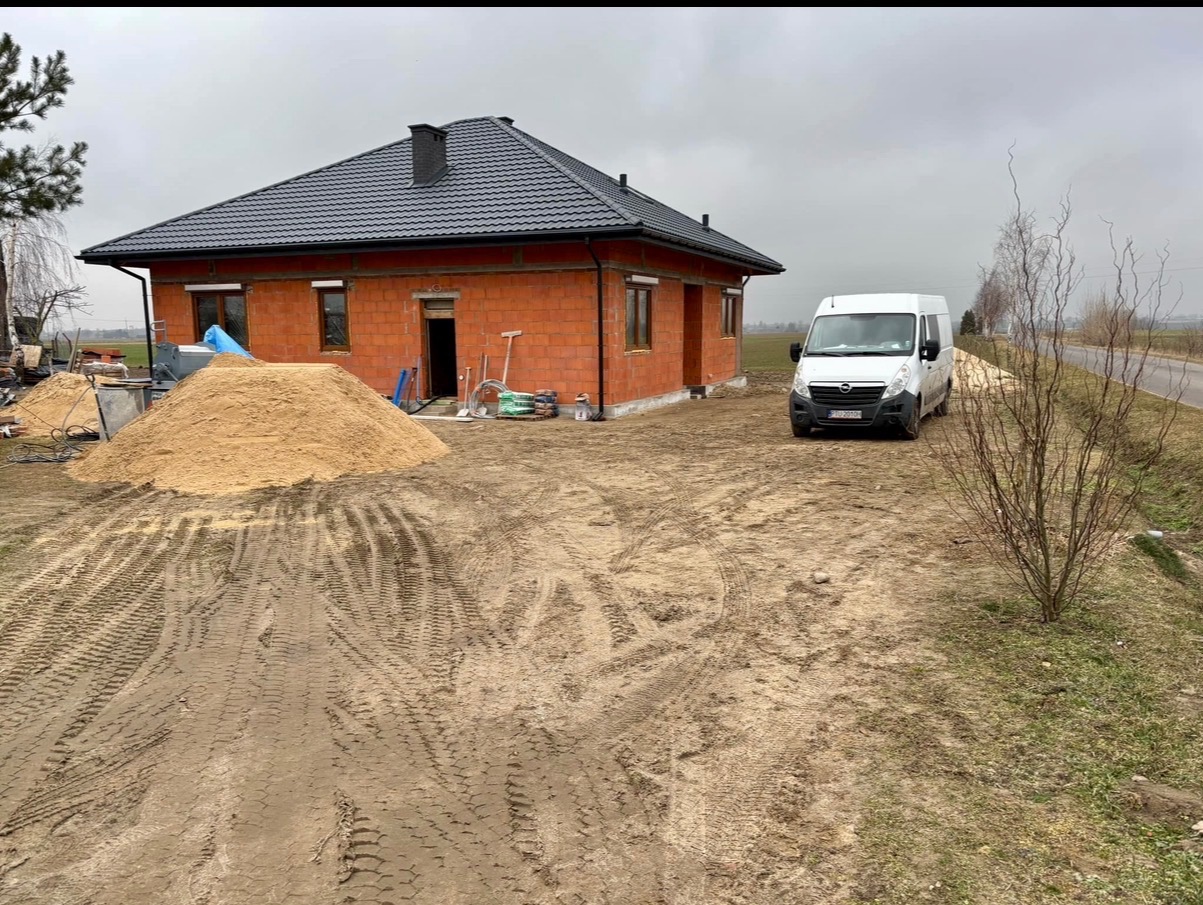 Dom w trakcie budowy z czerwonej cegły, z szarym dachem i stertą piasku obok. Biały bus zaparkowany na podwórku. Widok na pole w tle.