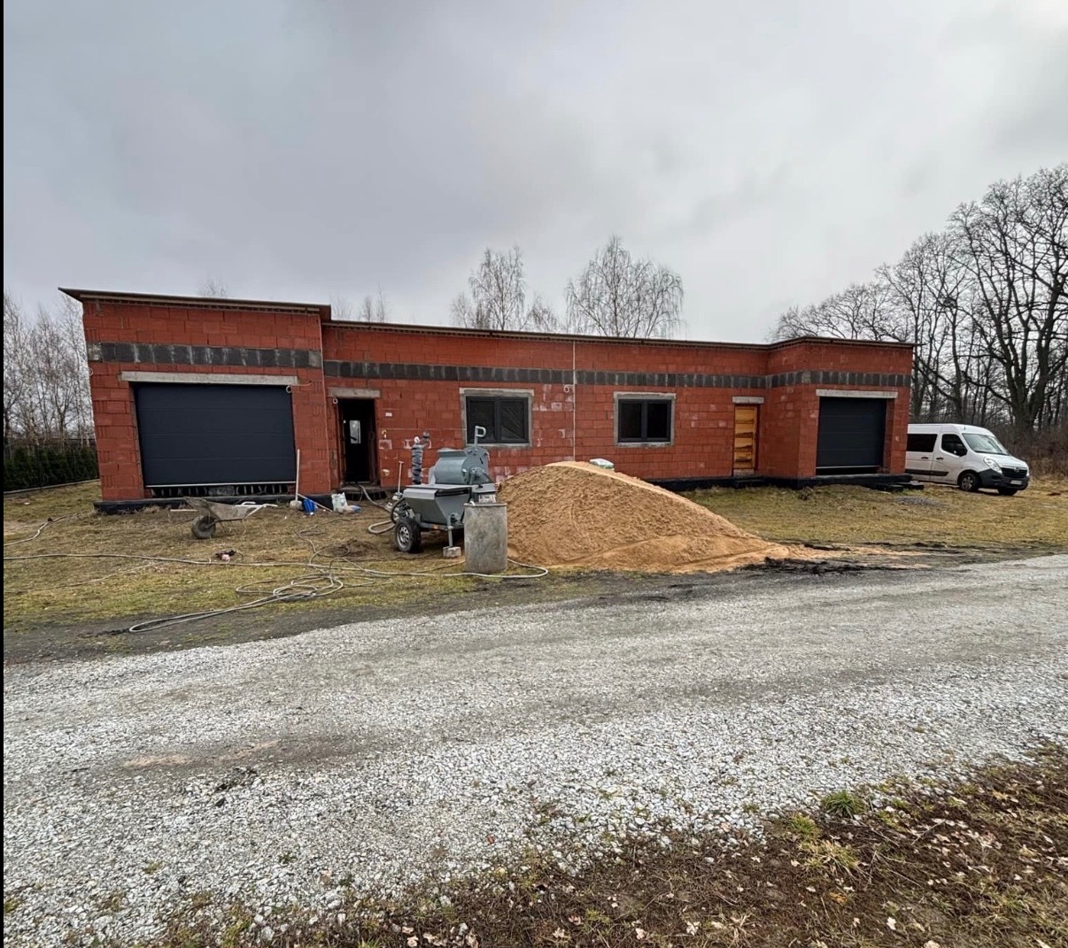 Budynek w trakcie tynkowania maszynowego, widoczna maszyna do tynkowania, piasek, surowe ściany z czerwonej cegły i brama garażowa. Na zewnątrz zaparkowany biały bus.