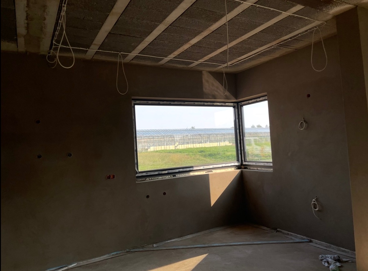 Pokój po nałożeniu tynku maszynowego, widoczne okna narożne z widokiem na szklarnie. Instalacja elektryczna na suficie. Świeżo otynkowane ściany w odcieniach szarości.
