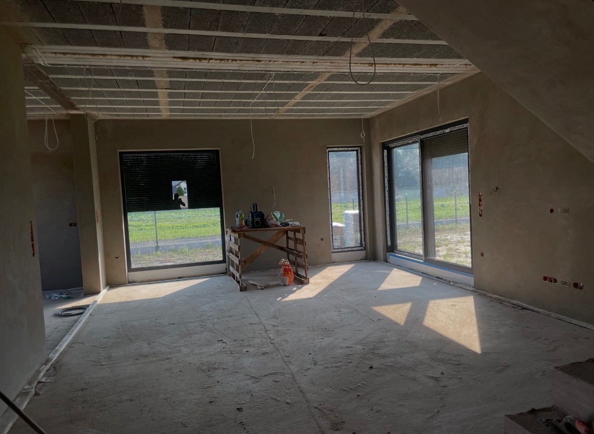 Surowe wnętrze po tynkowaniu: ściany pokryte szarym tynkiem, widoczne okna z roletami, drewniany stół z narzędziami. Podłoga betonowa, sufit z elementami konstrukcyjnymi.