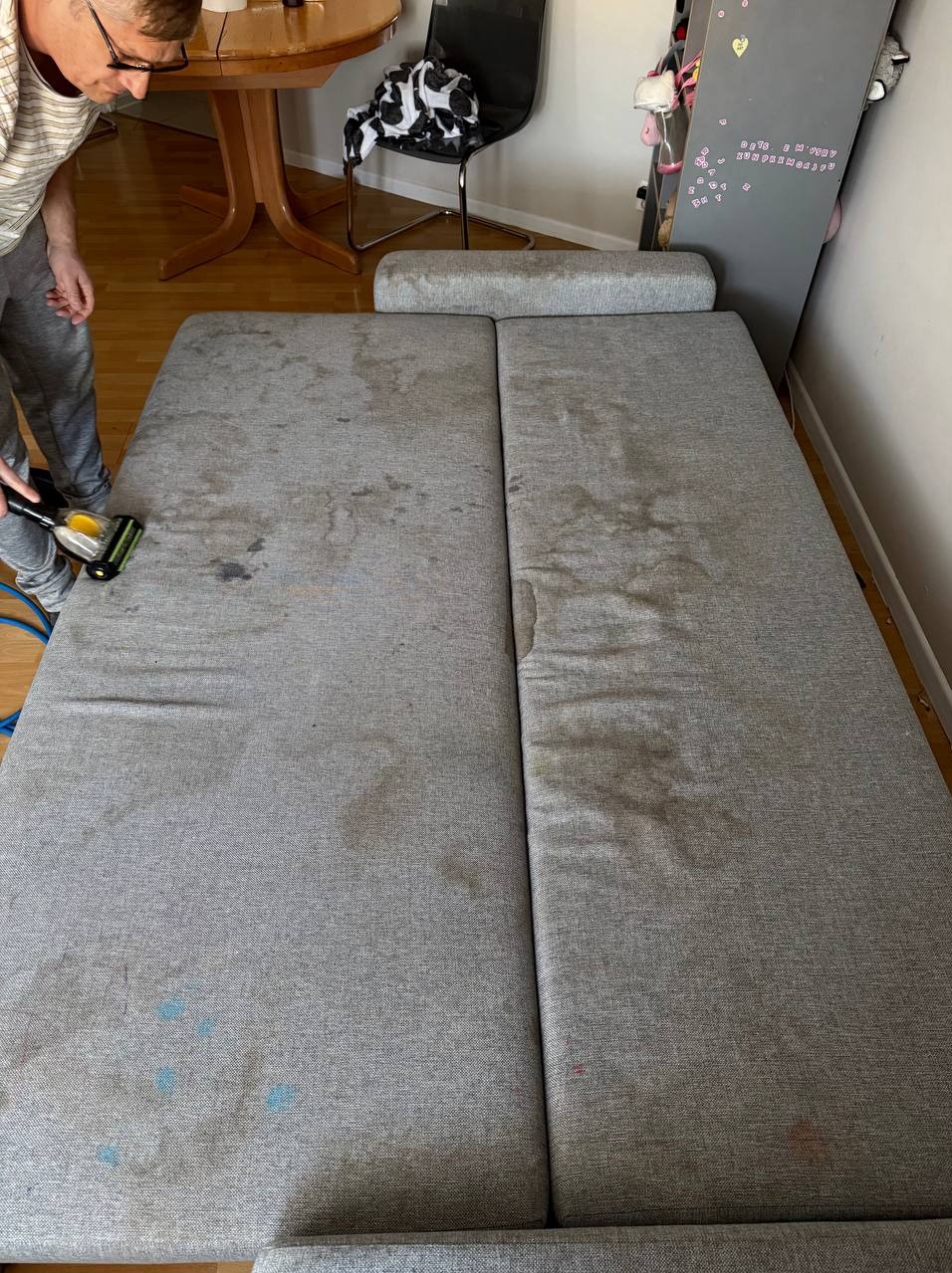 Czyszczenie sofy na mokro z widocznymi plamami. Osoba w trakcie pracy używa małego odkurzacza piorącego. Widoczne zabrudzenia na szarym materiale.