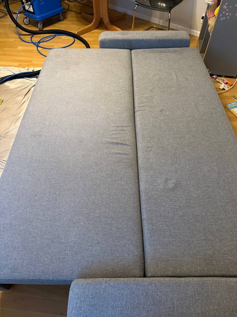 Szara, rozłożona sofa z widocznymi plamami po czyszczeniu, obok stoi niebieski odkurzacz piorący. Widok z góry, na drewnianej podłodze.