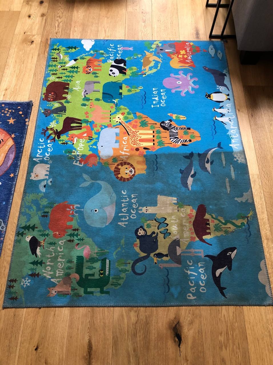 Kolorowy dywan dziecięcy z motywem mapy świata i zwierząt na drewnianej podłodze. Widoczny fragment drugiego dywanu z lewej strony.
