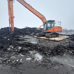 Maszyny-Long Mikołaj Milczarek - Pomarańczowa koparka Doosan na drewnianej platformie, pracująca na błotnistym terenie z resztkami śniegu. W tle widoczny inny sprzęt budowlany i budynek.