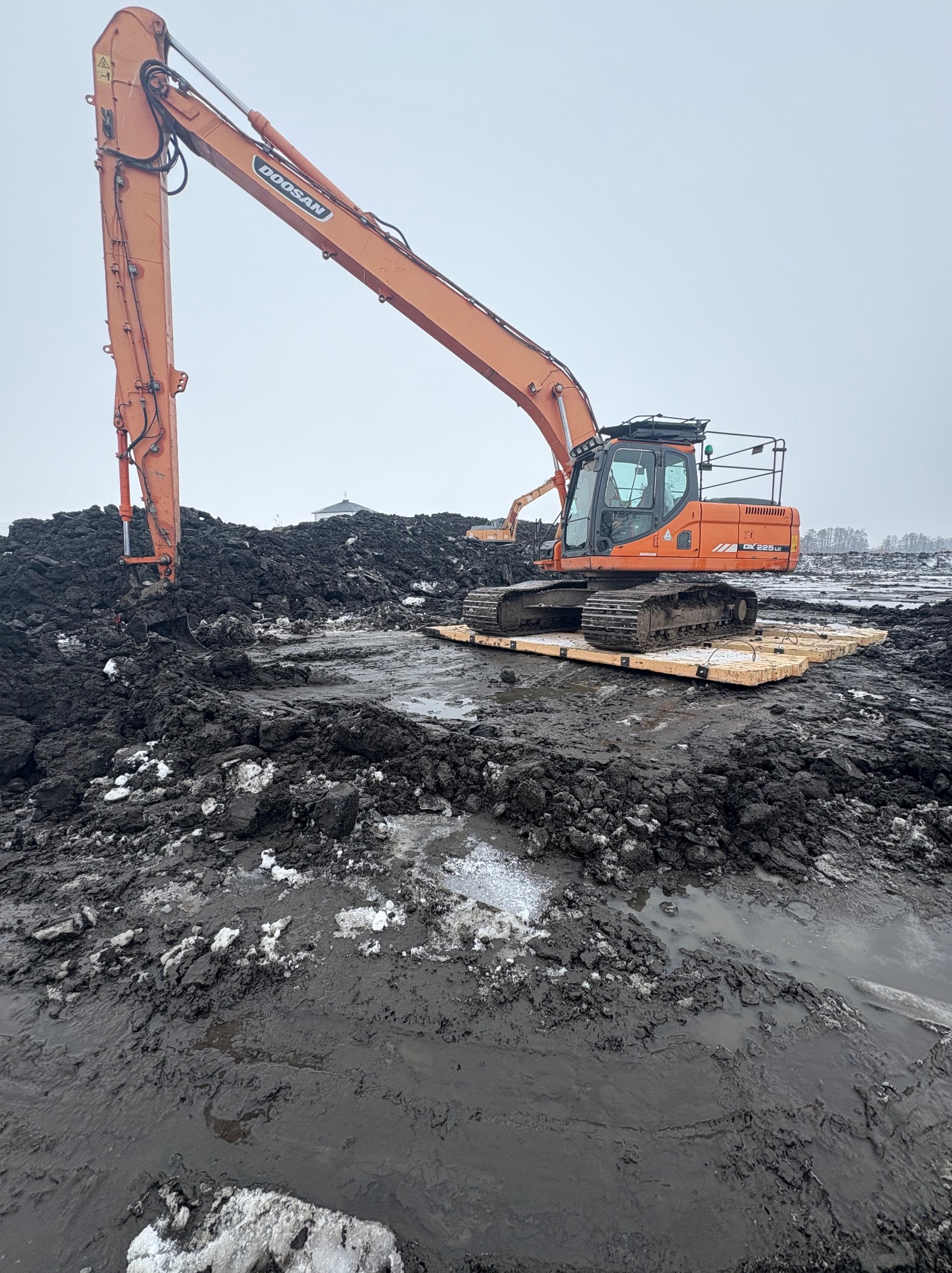 Pomarańczowa koparka Doosan na drewnianej platformie, pracująca na błotnistym terenie z resztkami śniegu. W tle widoczny inny sprzęt budowlany i budynek.