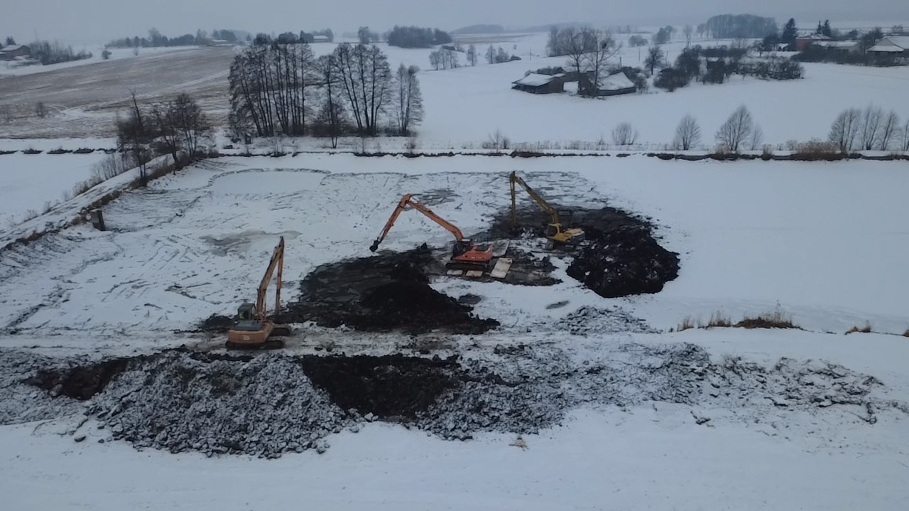 Zimowy krajobraz z trzema koparkami pracującymi przy wykopie stawu. Śnieg pokrywa teren, a w tle widać zabudowania wiejskie. Koparki w różnych fazach robót ziemnych.