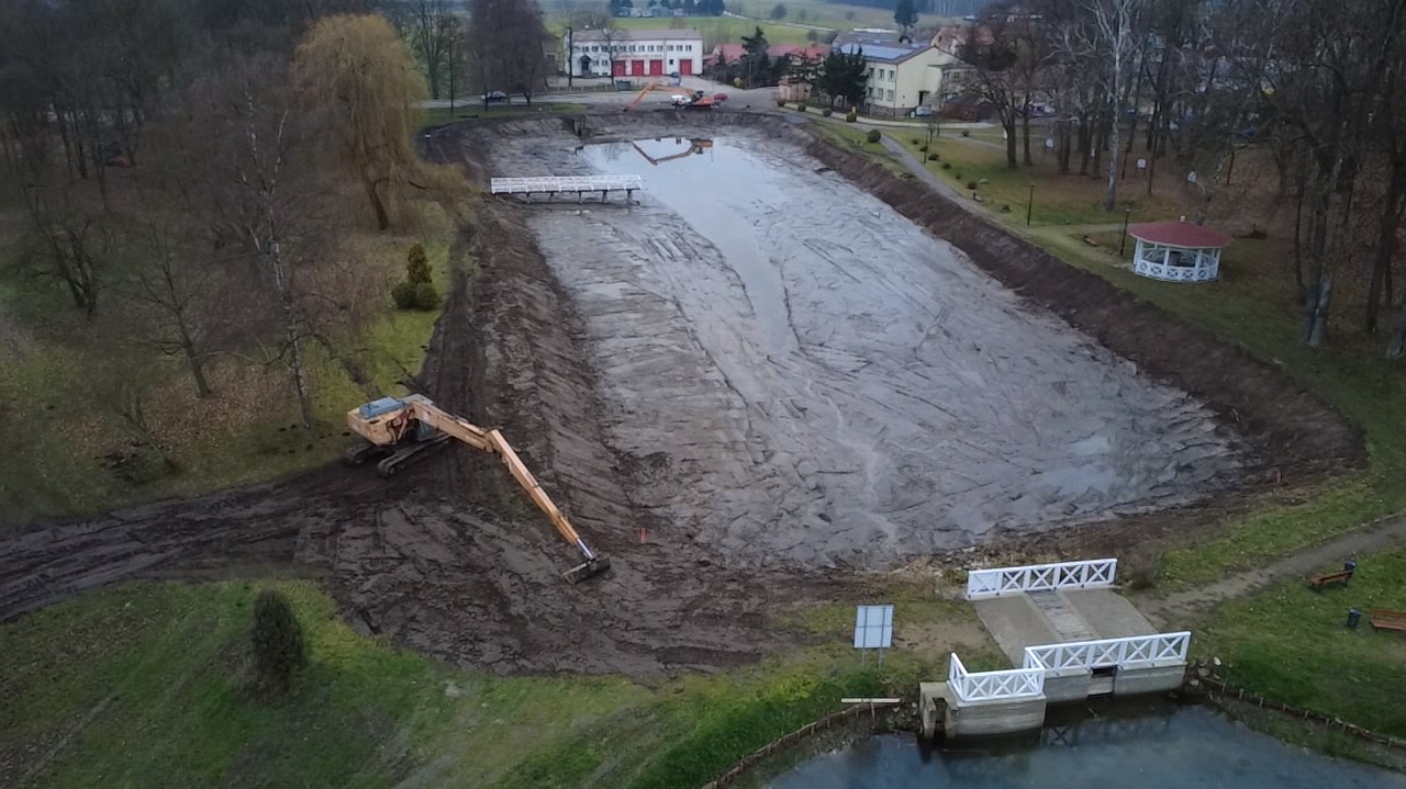 Prace ziemne przy stawie: koparka na brzegu, wykopany teren, widoczne pomosty i otoczenie parkowe. Ujęcie z góry ukazuje postęp prac.