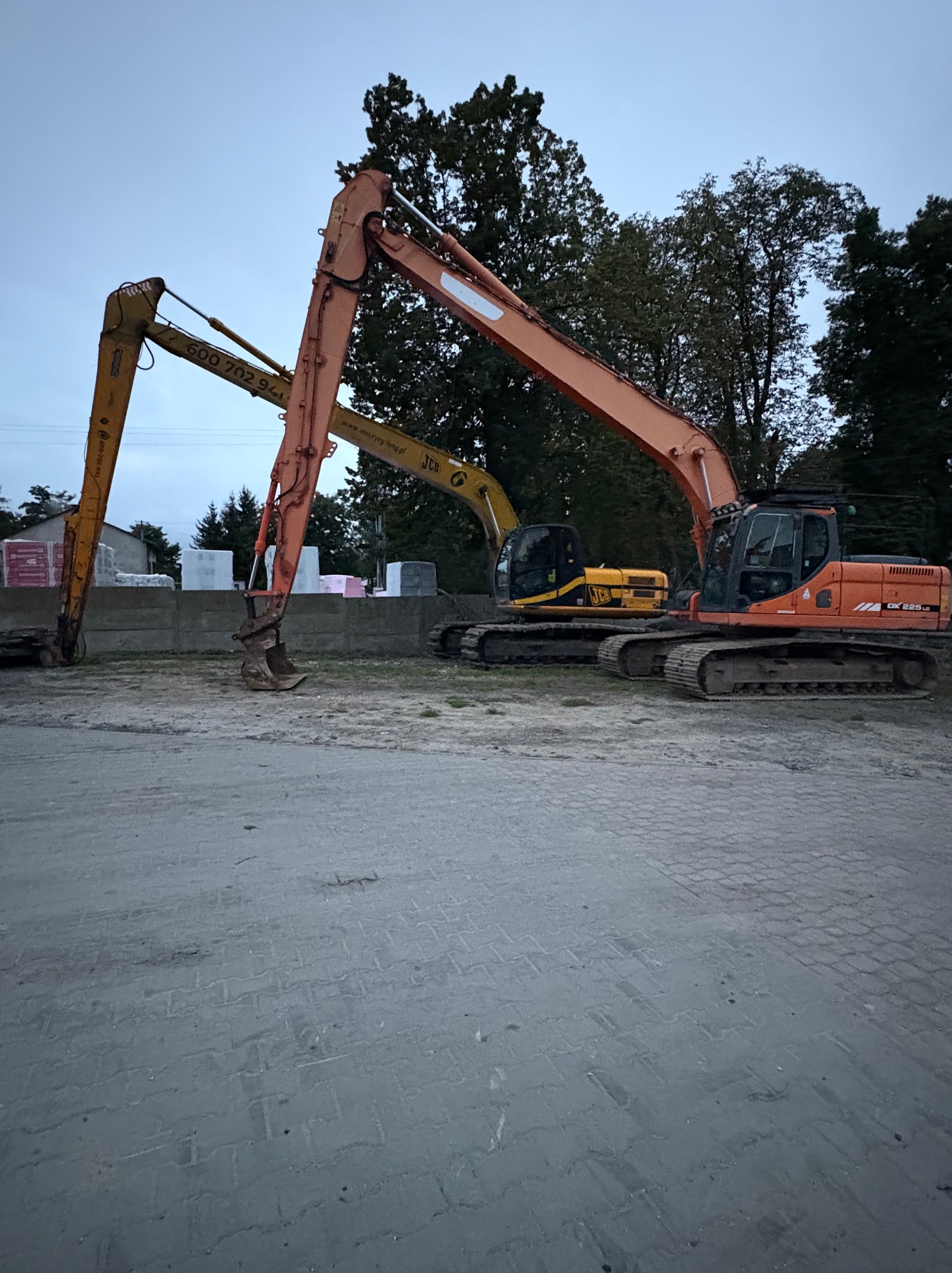 Dwa zaparkowane, pomarańczowo-żółte, ciężkie koparki na placu budowy, z podniesionymi ramionami i łyżkami, na tle drzew i materiałów budowlanych.