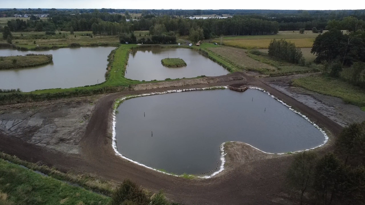 Widok z góry na kompleks stawów po wykopaniu, otoczonych zielenią i polami. Jeden ze stawów ma wyspę. Brzegi stawów zabezpieczone białym materiałem.
