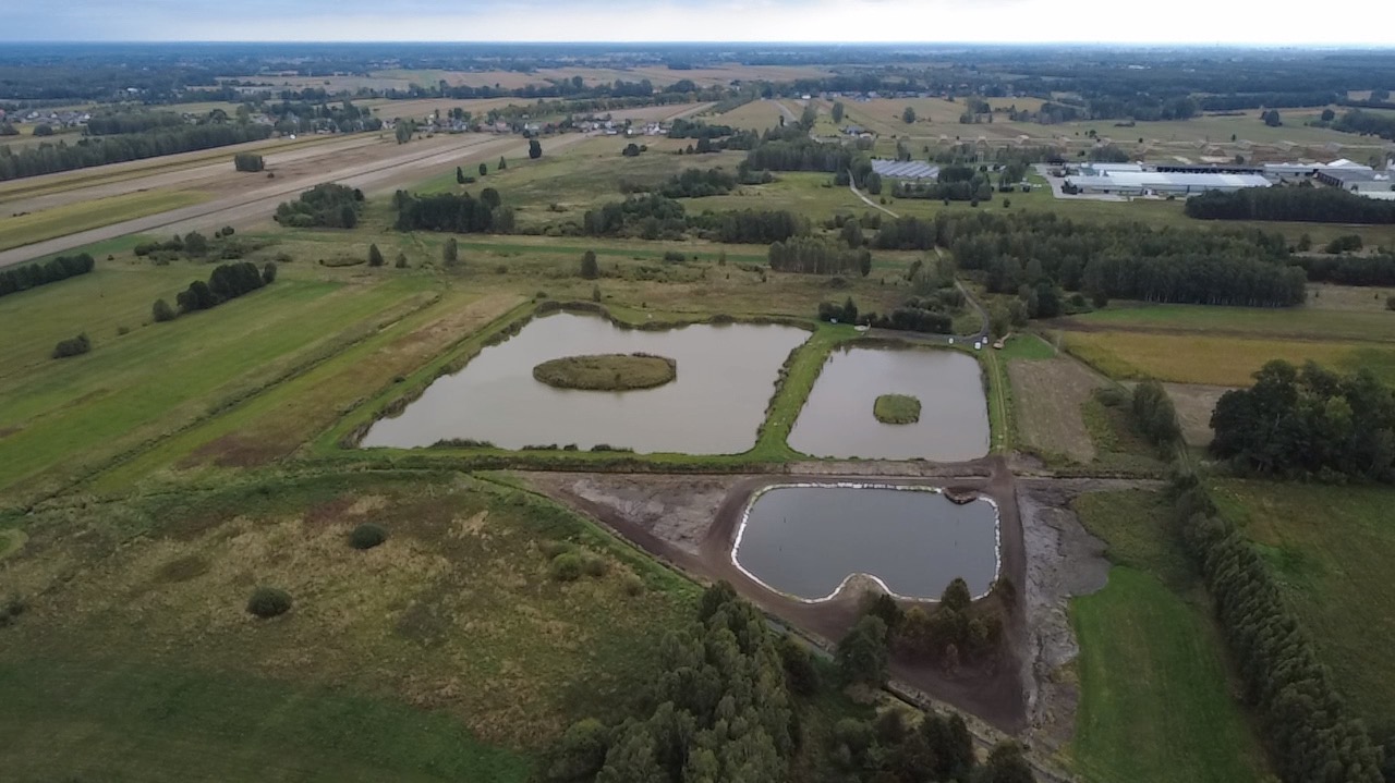 Widok z lotu ptaka na kompleks trzech stawów o różnych kształtach, z wyspami na dwóch z nich, otoczonych zielenią pól i lasów, w pochmurny dzień.