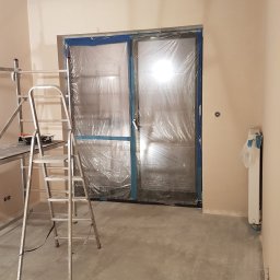 Idea Dom Usługi Remontowo-Wykończeniowe Wojciech Zakrzewski - Pokój w trakcie malowania: ściany w beżowym odcieniu, zabezpieczone folią drzwi, drabina malarska i taśma malarska przy suficie. Widoczny grzejnik i surowa podłoga.