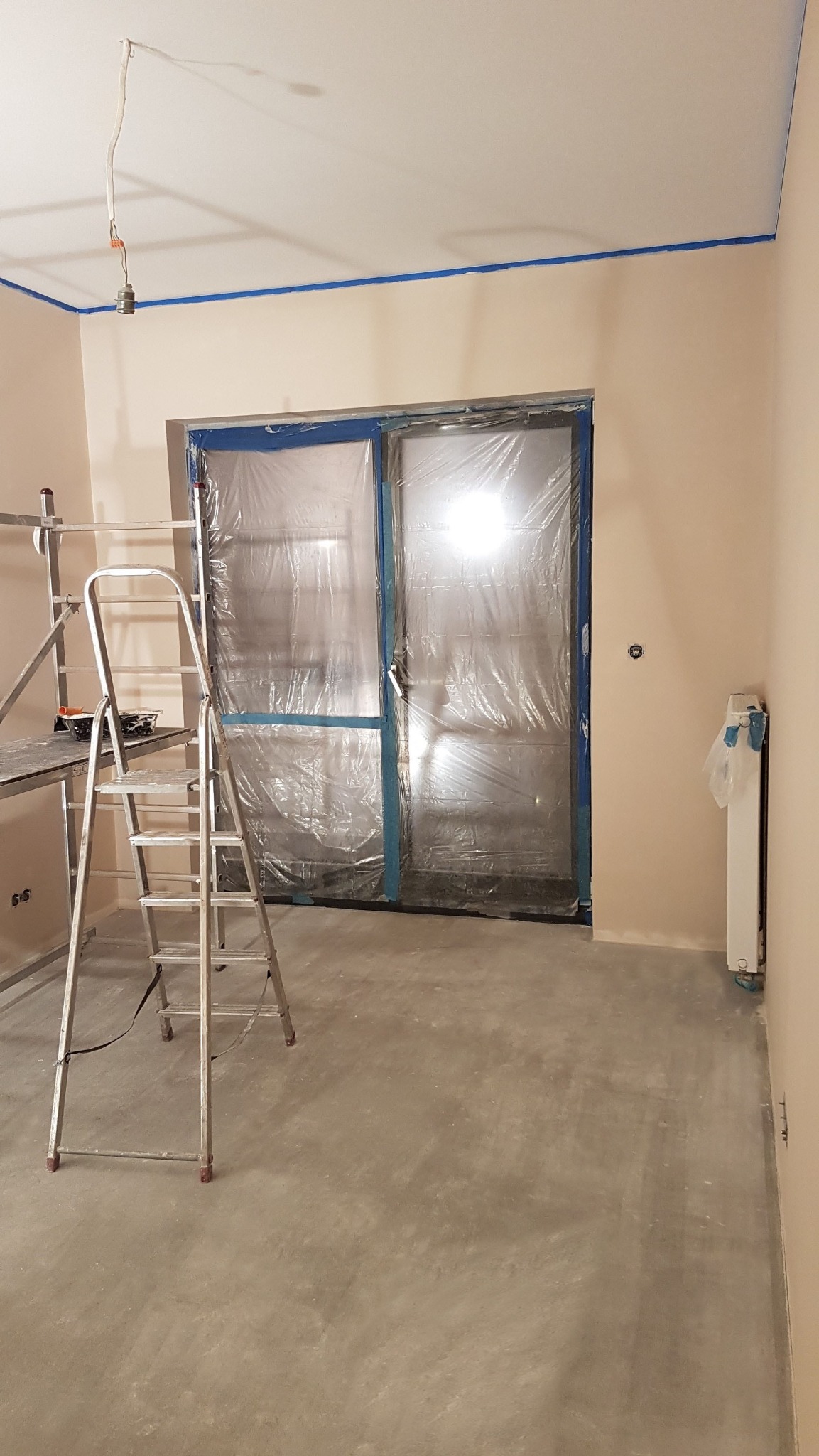 Pokój w trakcie malowania: ściany w beżowym odcieniu, zabezpieczone folią drzwi, drabina malarska i taśma malarska przy suficie. Widoczny grzejnik i surowa podłoga.