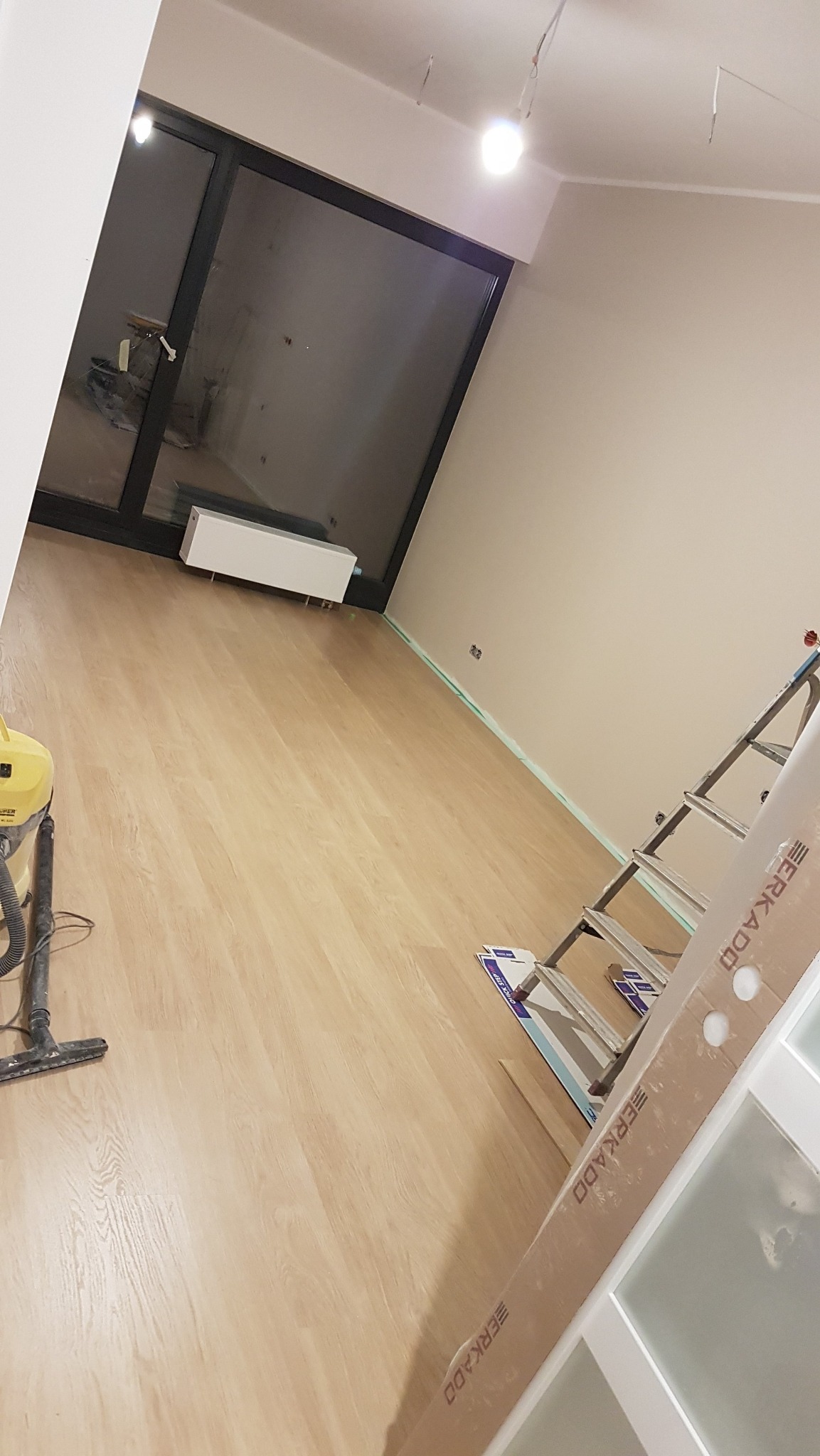 Jasne panele podłogowe w nowym pokoju z dużym oknem, drabina oparta o ścianę, odkurzacz i listwa przypodłogowa w tle. Widok na wnętrze w trakcie prac.