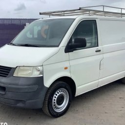 Prywatna - Biały bus Volkswagen Transporter T5 z relingami dachowymi, zaparkowany na zewnątrz. Widok z boku, lekko od przodu. Szare felgi i zderzak. Fioletowe ogrodzenie w tle.