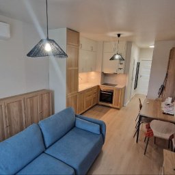 MISTRZOWIE BUDOWY SPÓŁKA Z OGRANICZONĄ ODPOWIEDZIALNOŚCIĄ - Jasne wnętrze mieszkania z aneksem kuchennym, niebieską sofą i drewnianymi szafkami. Widoczne lampy wiszące, stół z krzesłami i wzorzysta tapeta na ścianie.