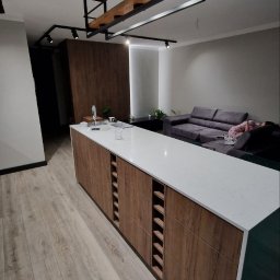 MISTRZOWIE BUDOWY SPÓŁKA Z OGRANICZONĄ ODPOWIEDZIALNOŚCIĄ - Nowoczesna wyspa kuchenna z drewnianymi frontami i białym blatem, oświetlona designerską lampą. Widoczny fragment salonu z szarą sofą i drewniana ściana.