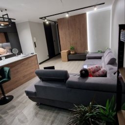 MISTRZOWIE BUDOWY SPÓŁKA Z OGRANICZONĄ ODPOWIEDZIALNOŚCIĄ - Nowoczesny salon z szarym narożnikiem, drewnianą ścianą akcentującą i oświetleniem szynowym. Jasna podłoga z paneli i rośliny doniczkowe dodają przytulności.