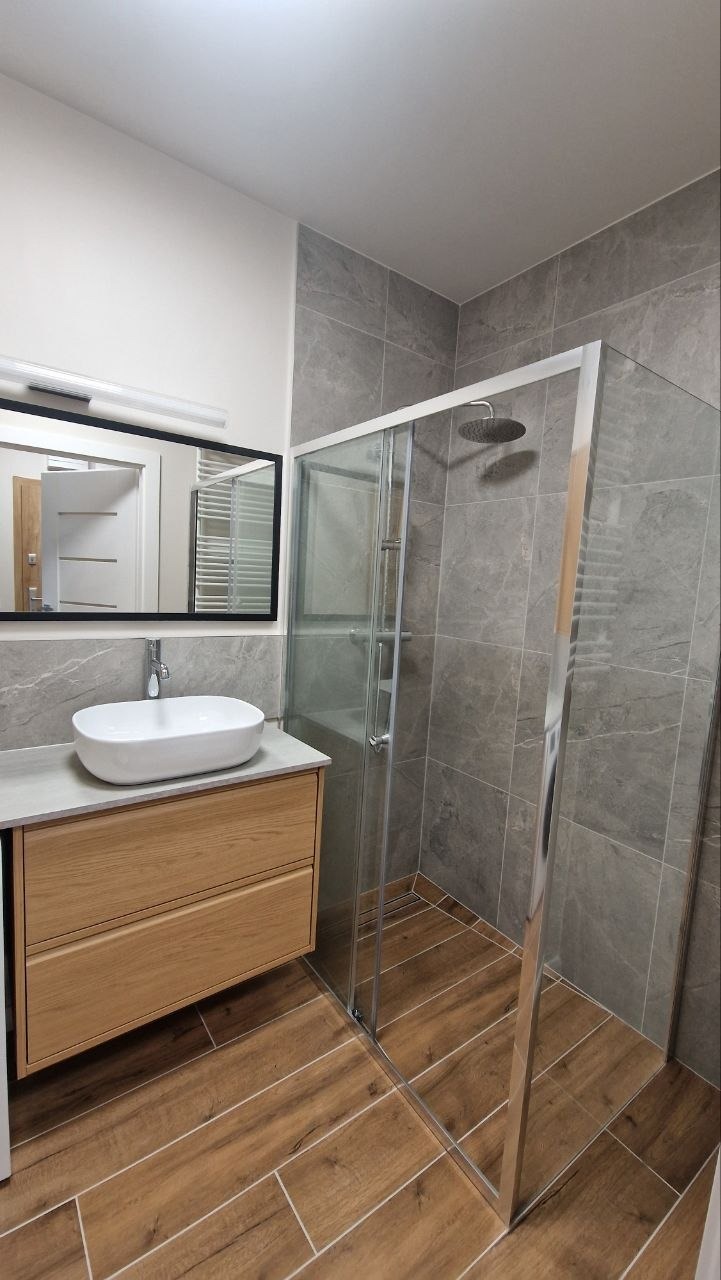 Nowoczesna łazienka z drewnianą szafką pod umywalką, szarą glazurą i kabiną prysznicową. Uwagę przykuwa drewnopodobna podłoga i minimalistyczne oświetlenie.