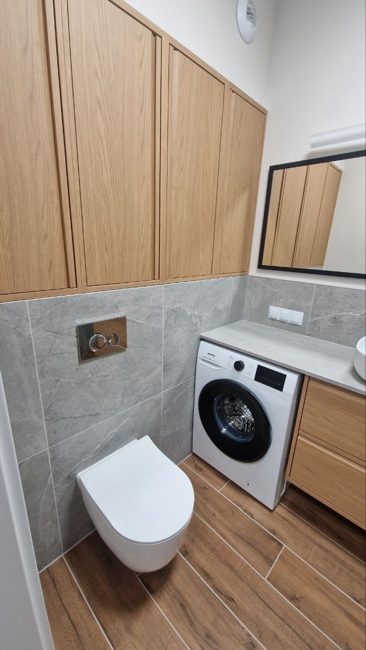 Nowoczesna łazienka po remoncie: podwieszana toaleta, pralka, drewniane szafki i płytki imitujące drewno na podłodze. Minimalistyczny design z chromowanymi akcentami.