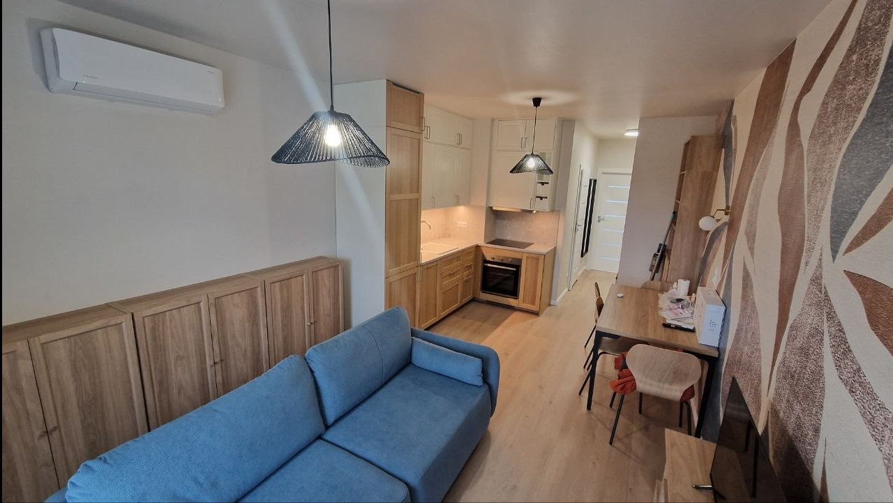 Jasne wnętrze mieszkania z aneksem kuchennym, niebieską sofą i drewnianymi szafkami. Widoczne lampy wiszące, stół z krzesłami i wzorzysta tapeta na ścianie.