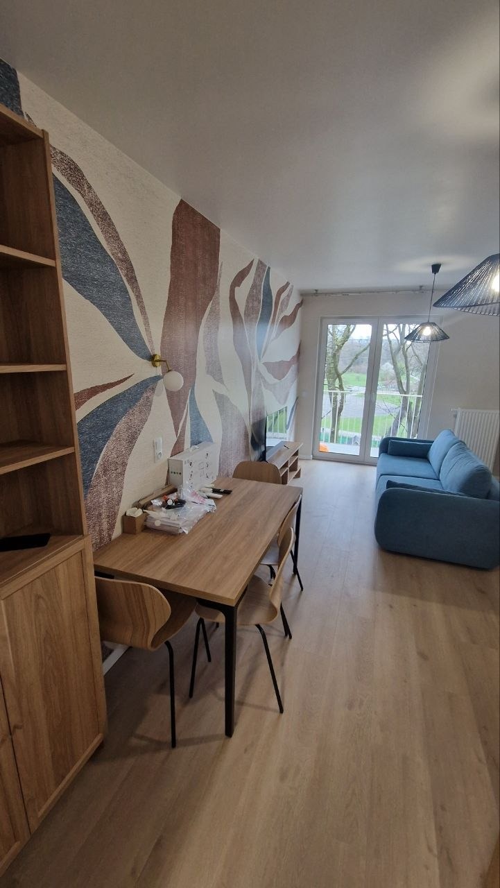 Jasne wnętrze salonu z drewnianą podłogą, stołem i krzesłami. Na ścianie dekoracyjna tapeta w abstrakcyjne wzory. Widoczny regał i niebieska sofa.