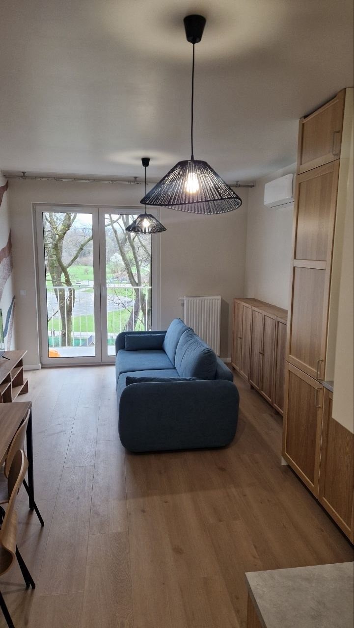 Jasne wnętrze z drewnianą podłogą, niebieską sofą i ażurowymi lampami. Widok na zieleń za oknem balkonowym. Szafki z jasnego drewna po prawej stronie.