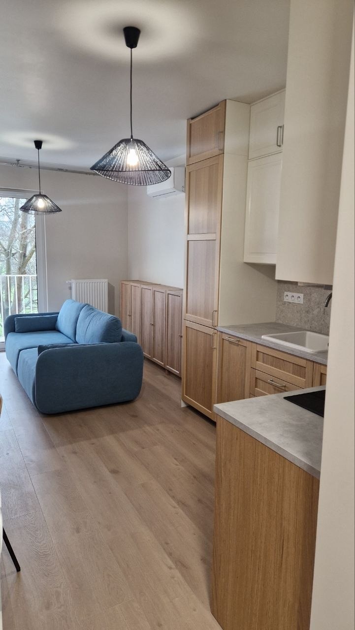 Jasne wnętrze po remoncie: parkiet, zabudowa kuchenna z drewnianymi frontami, niebieska sofa i lampy wiszące. Widok na balkon i drzewa za oknem.