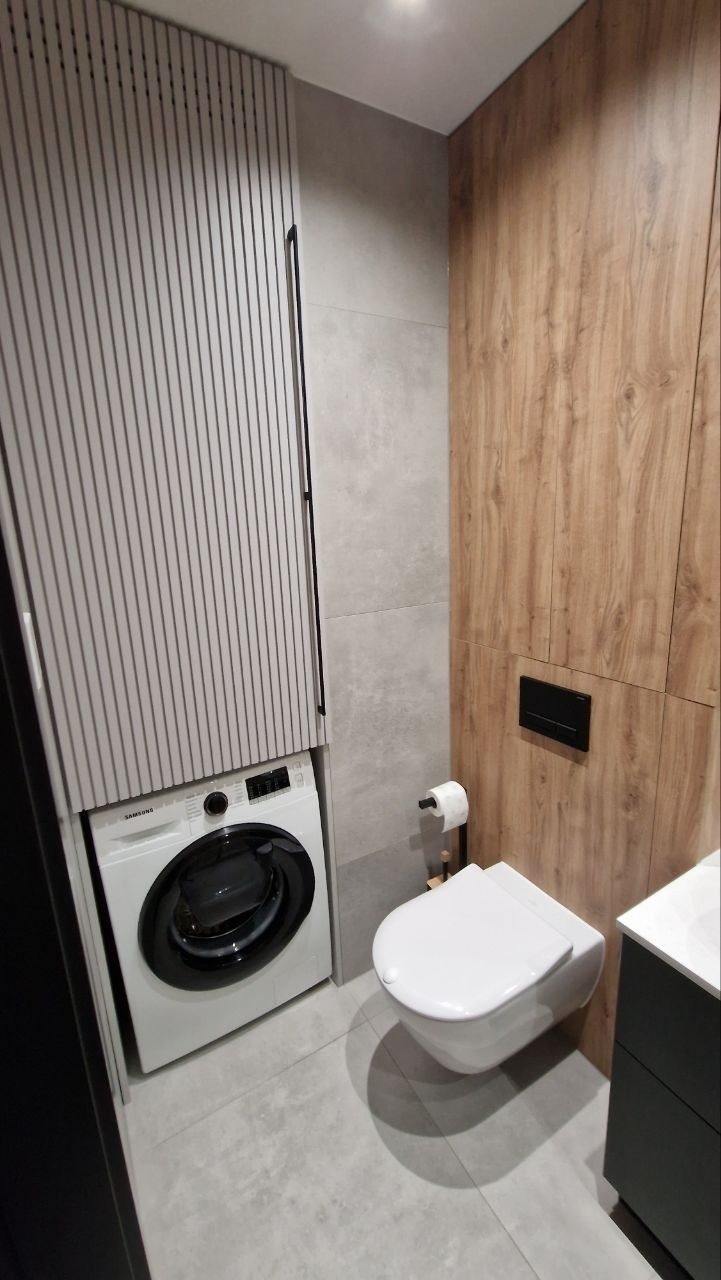 Nowoczesna łazienka z pralką ukrytą w zabudowie, wisząca toaleta, drewniane panele ścienne i szare płytki podłogowe. Minimalistyczny design.