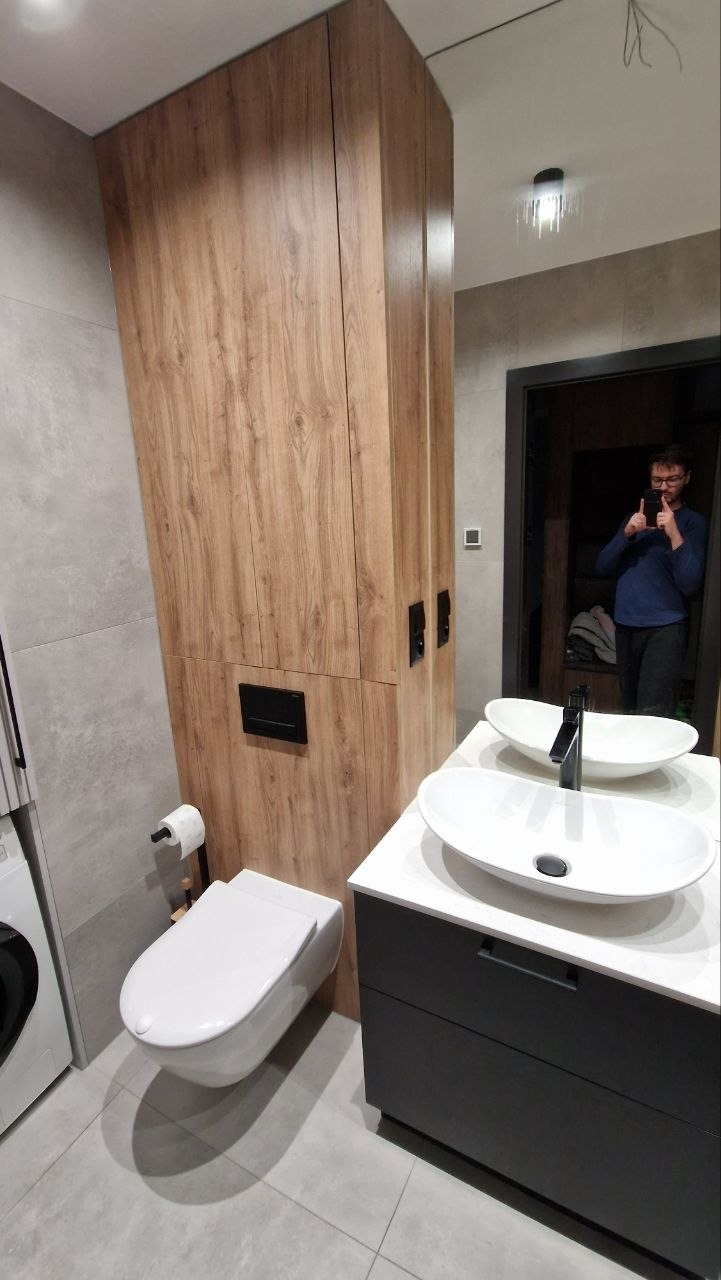 Nowoczesna łazienka z drewnianą zabudową, podwieszaną toaletą i umywalką nablatową. W lustrze odbija się osoba robiąca zdjęcie.