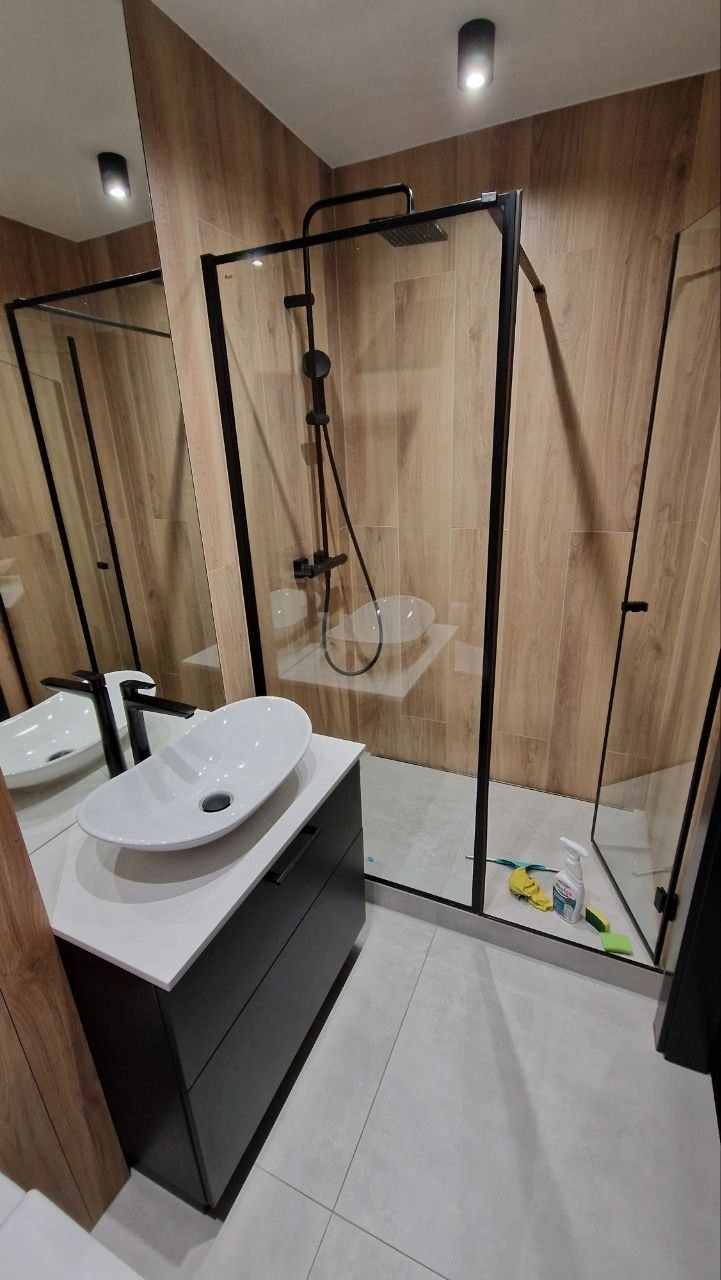 Nowoczesna łazienka z kabiną prysznicową typu walk-in z czarnymi profilami, umywalką nablatową i drewnopodobnymi płytkami ściennymi. Minimalistyczny design.