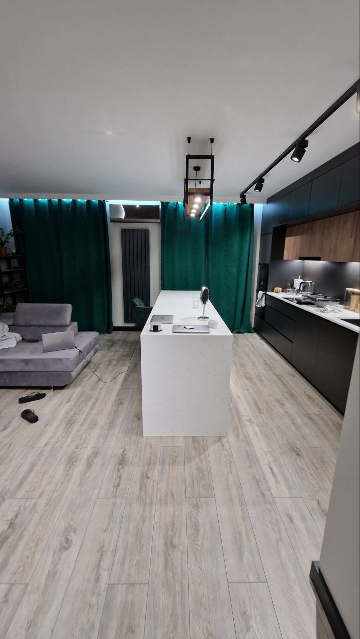 Nowoczesna kuchnia z wyspą i drewnianymi akcentami, oświetlenie szynowe, podłoga imitująca drewno, zielone zasłony w tle. Widok na salon z szarą sofą.