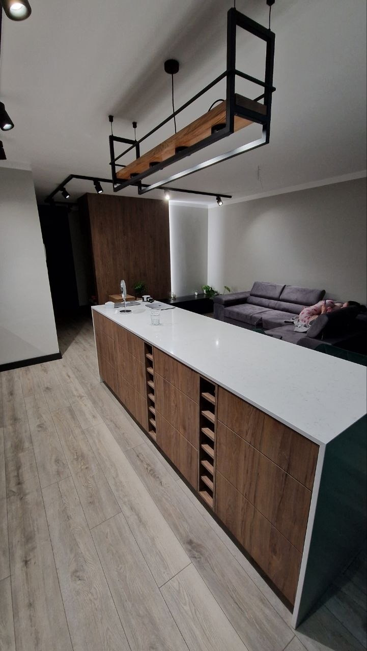 Nowoczesna wyspa kuchenna z drewnianymi frontami i białym blatem, oświetlona designerską lampą. Widoczny fragment salonu z szarą sofą i drewniana ściana.
