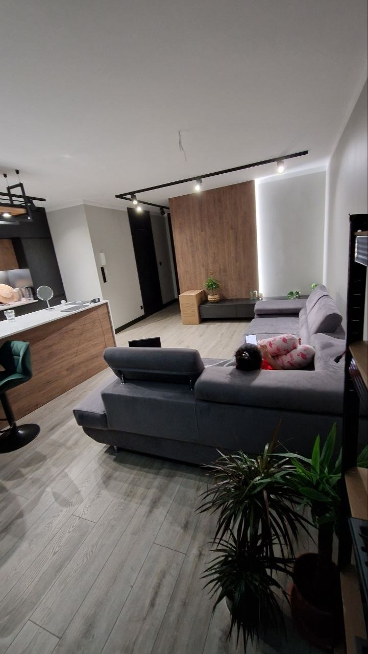 Nowoczesny salon z szarym narożnikiem, drewnianą ścianą akcentującą i oświetleniem szynowym. Jasna podłoga z paneli i rośliny doniczkowe dodają przytulności.