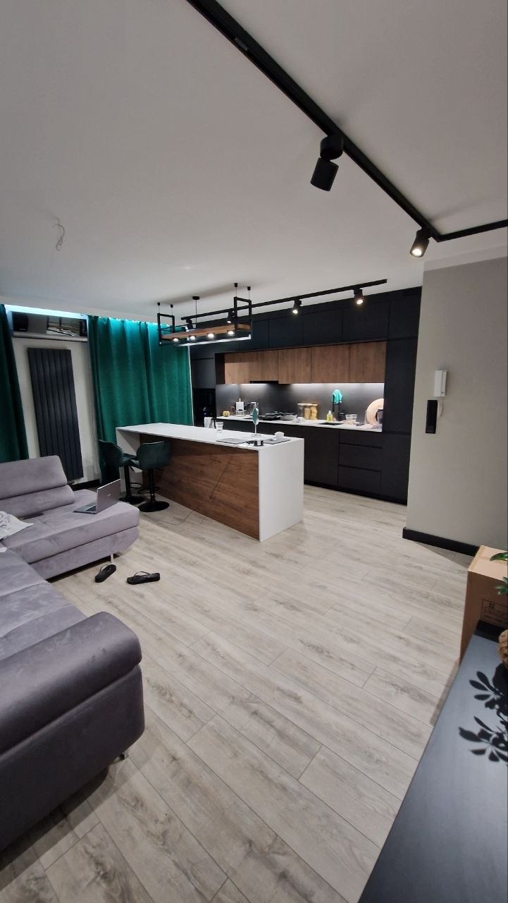 Nowoczesna kuchnia z wyspą, drewniane i czarne szafki, oświetlenie punktowe na szynoprzewodzie, szare panele podłogowe, widok z salonu.