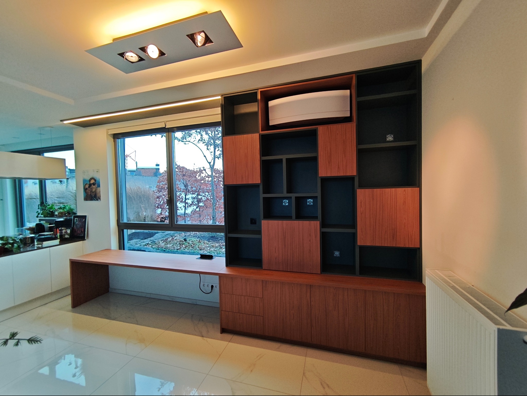 Nowoczesna zabudowa meblowa z biurkiem w salonie, drewniane fronty, czarne półki, oświetlenie sufitowe, widok na zewnątrz przez okno. Minimalistyczny design.