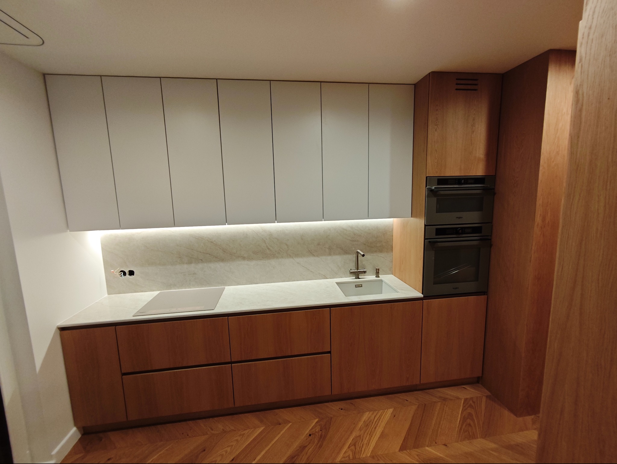 Nowoczesna kuchnia z drewnianymi szafkami dolnymi i białymi górnymi, marmurowym blatem i podświetleniem LED. Parkiet w jodełkę na podłodze.