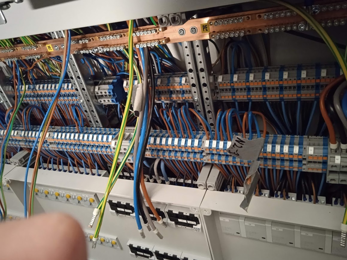 Rozdzielnia elektryczna z licznymi przewodami w kolorach: niebieskim, brązowym, zielono-żółtym. Widoczne szyny PE i N. Elementy metalowe i plastikowe.