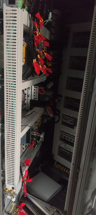 Rozdzielnica elektryczna z przewodami oznaczonymi czerwonymi zaciskami, widok z boku, ukazujący skomplikowaną sieć połączeń i elementów sterujących.