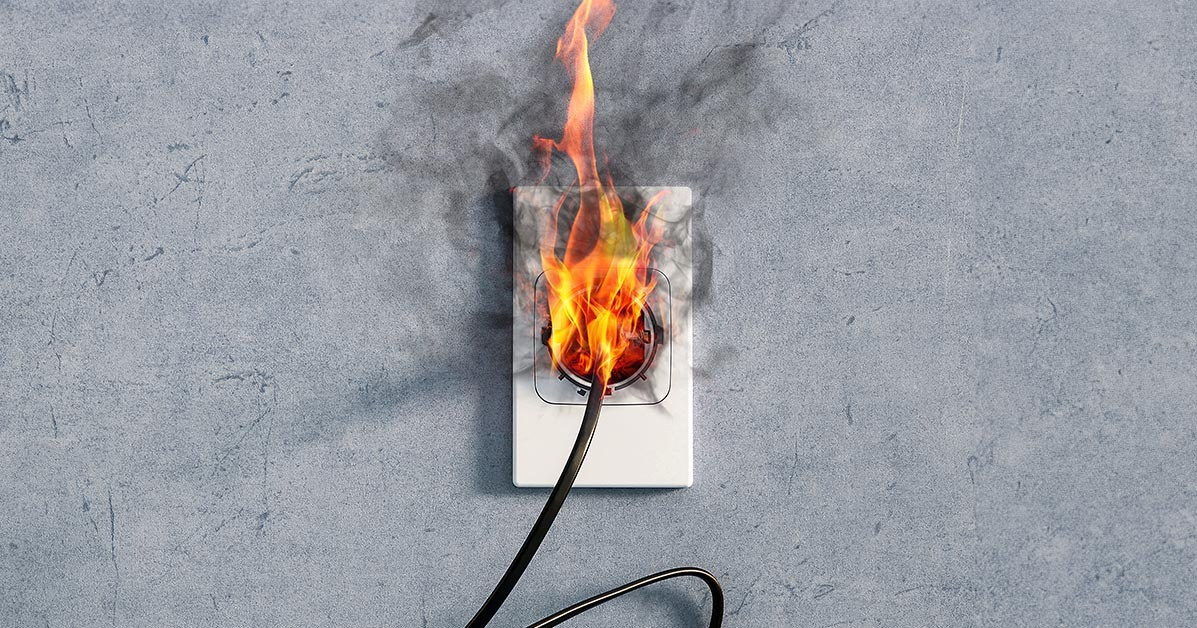 Płonące gniazdko elektryczne z czarnym kablem na szarej ścianie. Ogień i dym wydobywają się z gniazdka, sugerując awarię lub zwarcie.