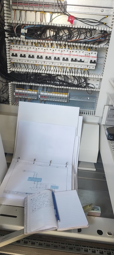 Rozdzielnica elektryczna z widocznymi bezpiecznikami, przewodami i schematem w segregatorze. Notatnik z długopisem leżą obok, sugerując prace instalacyjne.