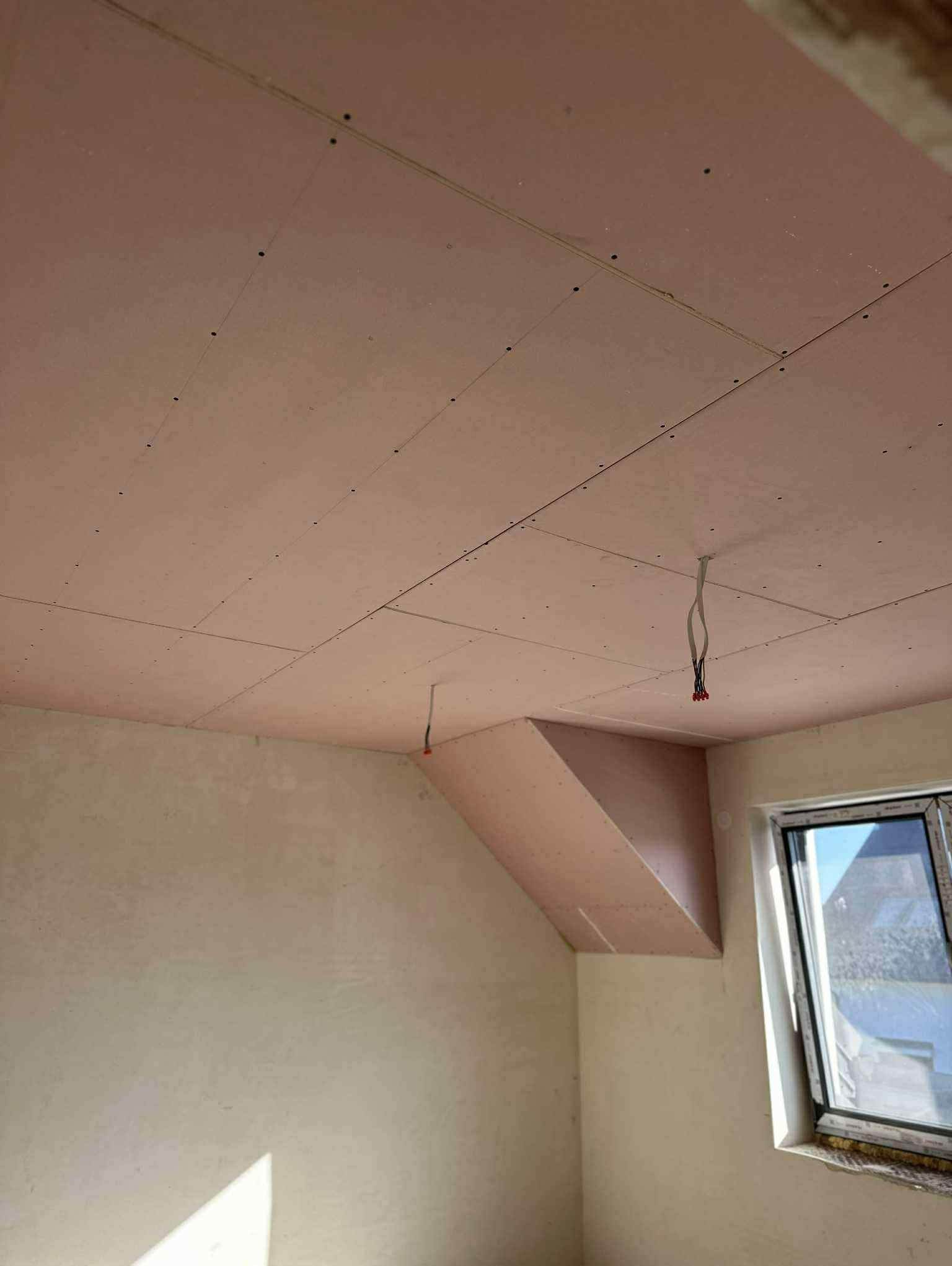 Różowa płyta gipsowo-kartonowa zamontowana na skośnym suficie poddasza, widoczne kable elektryczne i okno. Etap prac wykończeniowych.