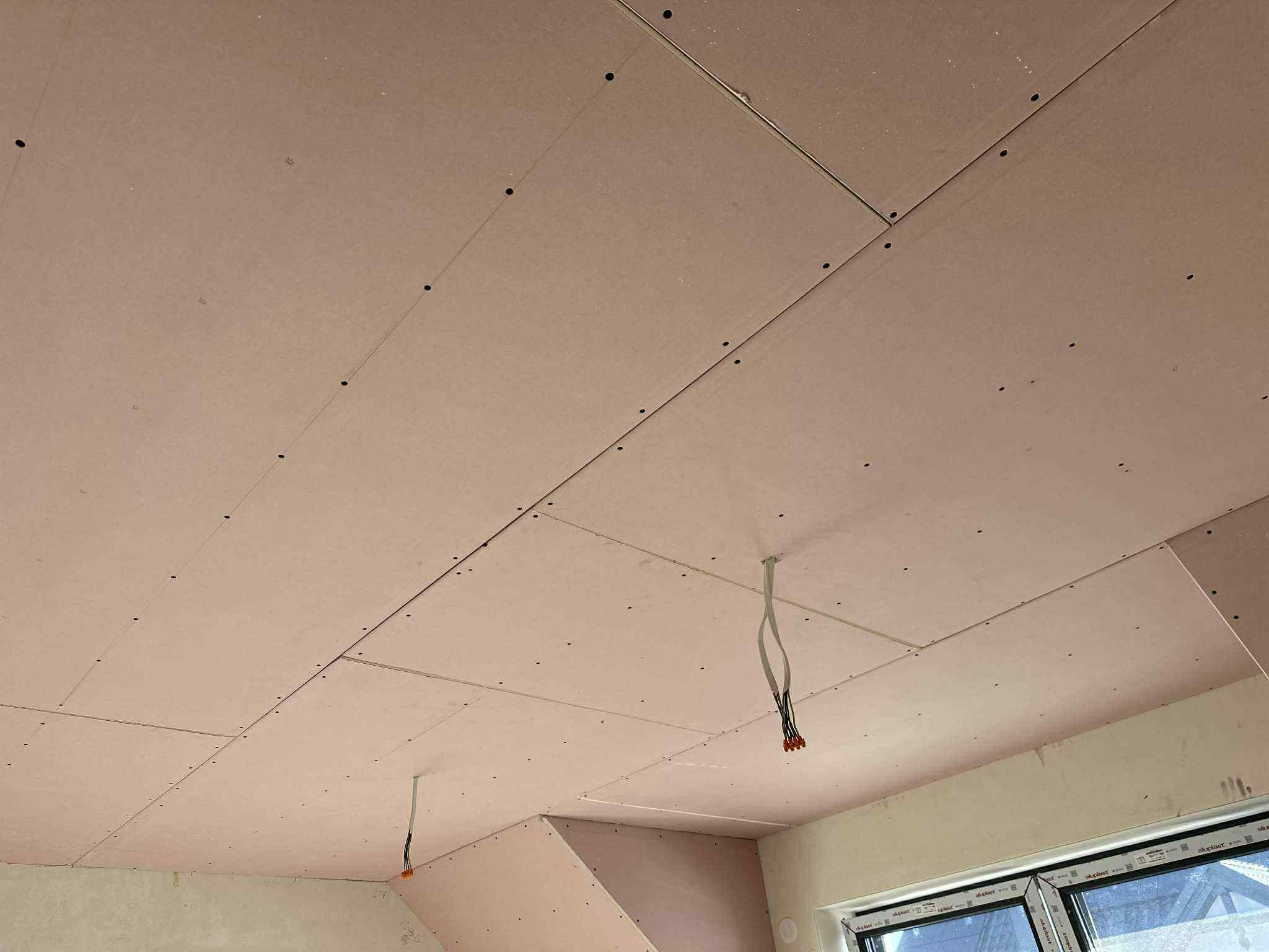 Różowe płyty karton-gips na skośnym poddaszu, widoczne łączenia i kable zwisające z sufitu. Okno w dolnej części kadru.