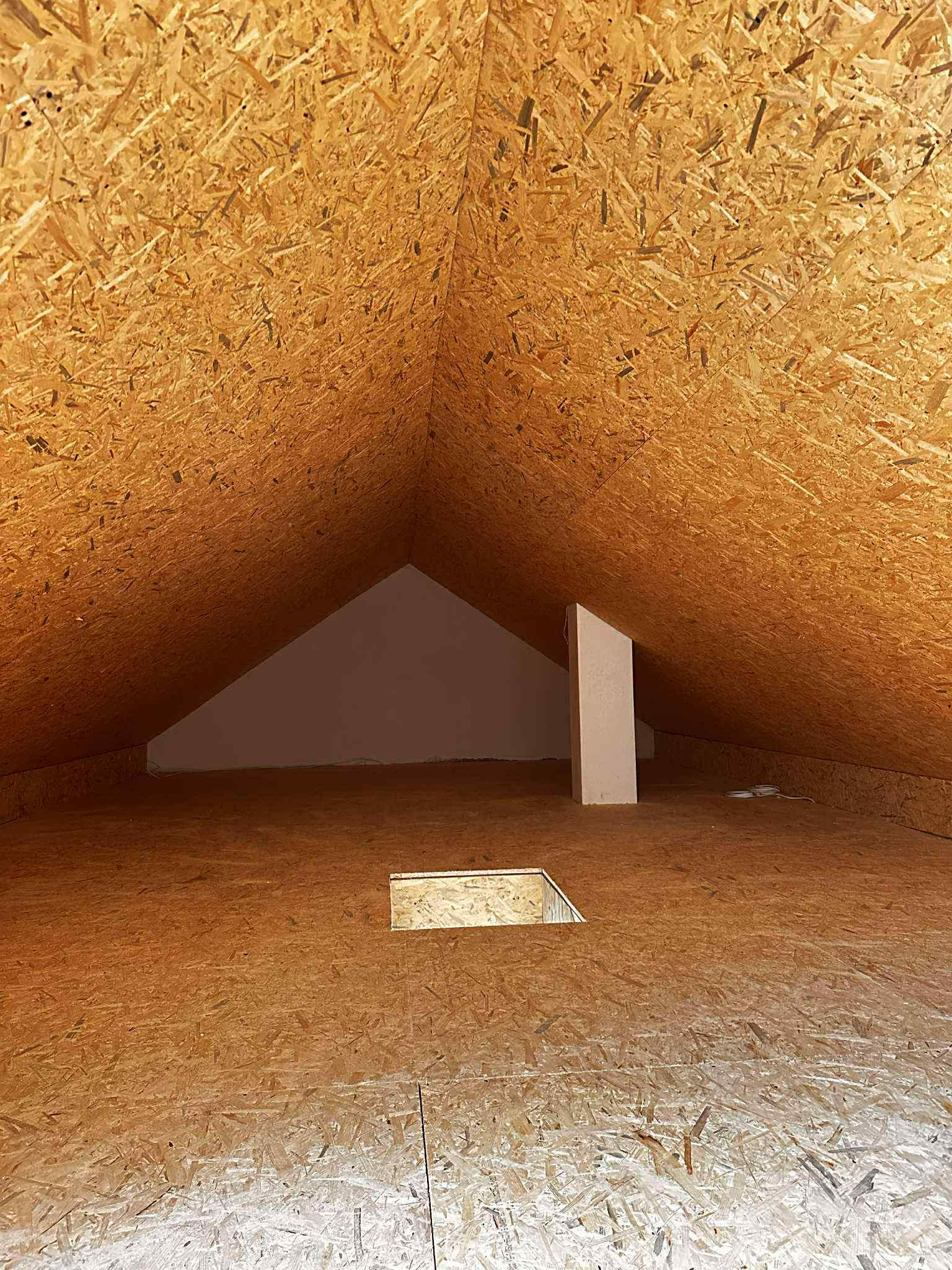 Poddasze wykończone płytami OSB, widoczny otwór w podłodze i pionowy element konstrukcyjny. Surowe wykończenie, przygotowanie do dalszych prac.