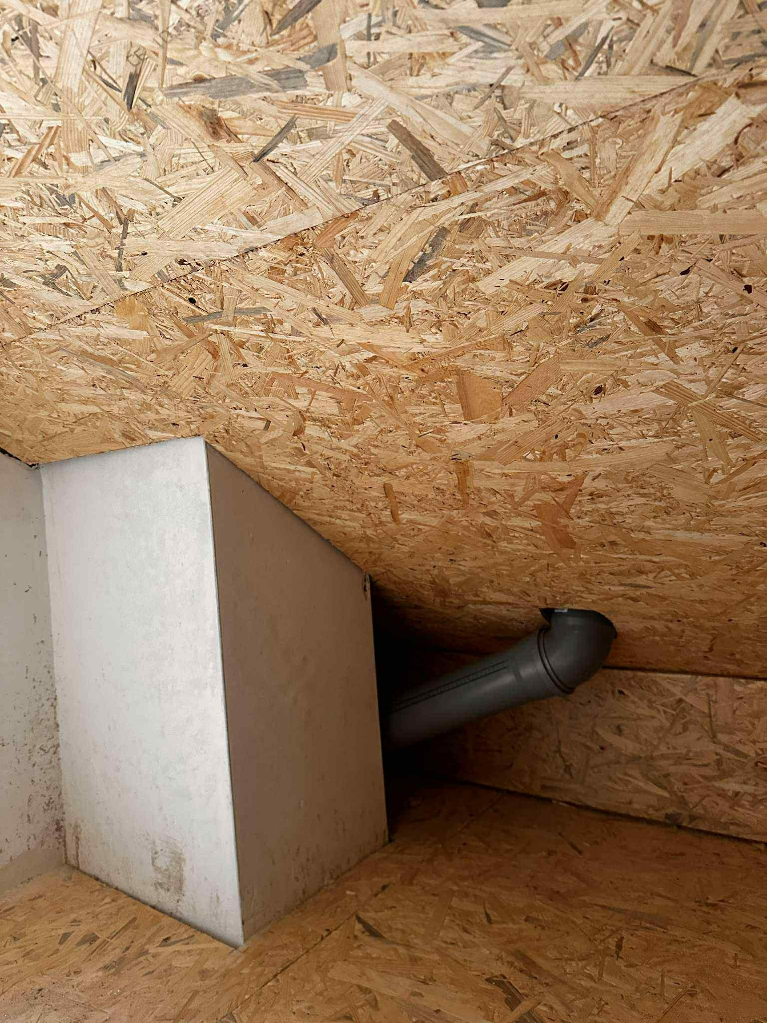 Poddasze z płyt OSB, fragment instalacji wentylacyjnej z szarą rurą i biała zabudowa. Widok z dołu, oświetlenie naturalne.