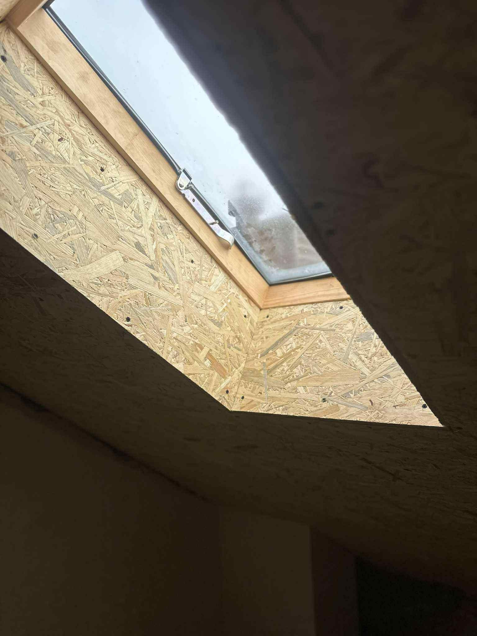 Montaż okna dachowego w poddaszu wykończonym płytą OSB. Widoczny fragment okna z metalowym uchwytem i drewniana rama. Ujęcie z dołu, perspektywa z wnętrza.