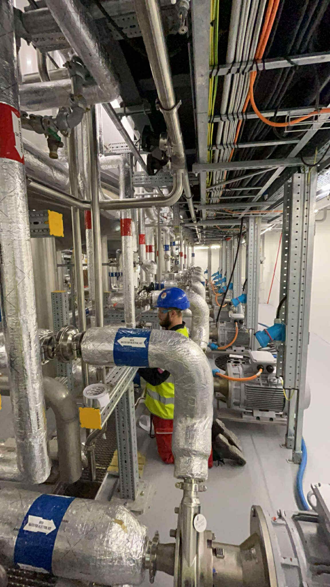 Pracownik w kasku i kamizelce odblaskowej izoluje rury srebrną folią. Widoczne liczne rury, kable i silniki. Instalacja przemysłowa w jasnym pomieszczeniu.