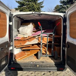 Usługi transportowe - Otwarty bagażnik białego vana wypełniony meblami i workiem, w tle drzewa. W bagażniku wózek transportowy z czerwonymi uchwytami. Widok z zewnątrz na kostkę brukową.