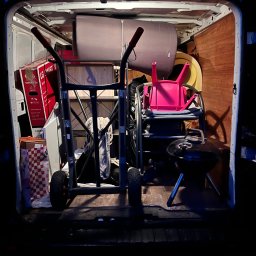 Usługi transportowe - Wnętrze busa załadowane różnorodnymi przedmiotami, w tym kartonami, meblami i grillem, przygotowanymi do transportu. Widoczny wózek transportowy.