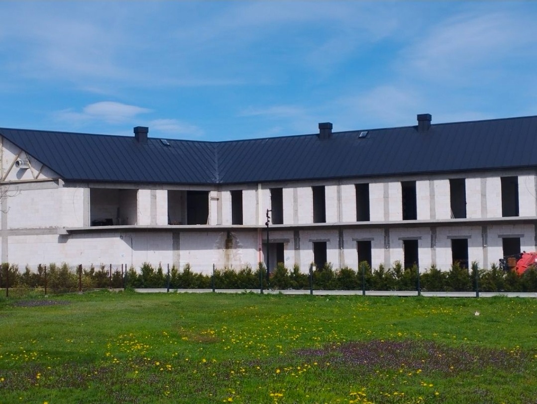 Budynek w stanie surowym zamkniętym z ciemnym dachem w Warszawie, otoczony zielenią. Widoczne niezadaszone otwory okienne i drzwiowe.